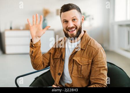 Cheerful Bearded Man Waving Hand Gesturing Hello Sitting Indoor