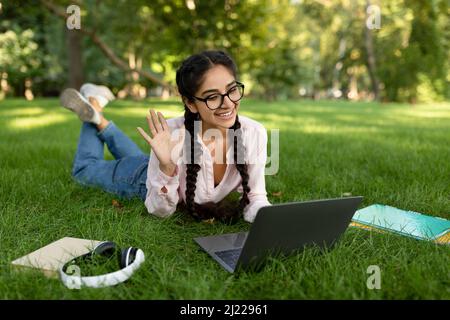 Cheerful female student waving hand to laptop while video calling, learning online, lying on lawn in park Stock Photo