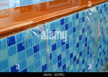 Closeup overflowing water of golden brown and vibrant blue tiled swimming pool spillway Stock Photo