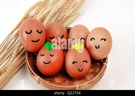 Indoor photography of bunch of eggs in a wicker basket with smiling faces on a white background with some dry wheat plants Stock Photo