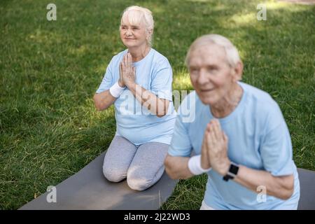 Smiling senior woman with husband do namaste practicing yoga on lawn grass in park Stock Photo