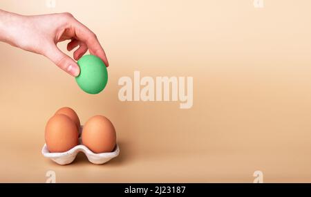 Banner with hand putting outstanding Easter egg in carton box with natural ones on beige background. Green symbol of religious holiday. Standing out from crowd concept. Copy space. High quality photo Stock Photo
