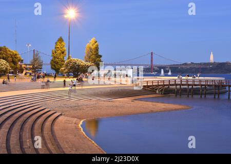 Tourists on the bank of river Tagus at dusk and modern 25 de Abril Bridge in background, Belem Lisbon Portugal. Stock Photo