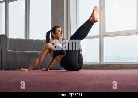 Side view of sporty girl exercising, sitting with raised legs indoors. Young brunette female practicing yoga, meditating alone, wearing black sports suit, Concept of yoga. Stock Photo