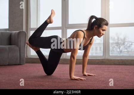 Side view of flexible girl exercising yoga pose indoors. Young slim female wearing black sports suit, standing on knee with raised bent leg, leaning on hands. Concept of yoga. Stock Photo