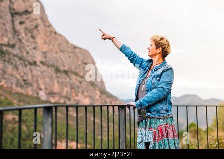 Adult woman, blonde, short hair, looking and pointing at a mountain view from a lookout point, on holiday. Stock Photo