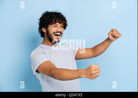 Joyful attractive curly haired indian or arabian guy, wearing casual t-shirt, holding in hands driving invisible car, imaginary steering wheel, stands on isolated blue background, smiling Stock Photo