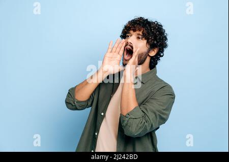 Excited indian or arabic curly haired guy in casual clothes, shouting loudly holding his hands near his mouth, making an announcement, calling out, standing on an isolated blue background Stock Photo