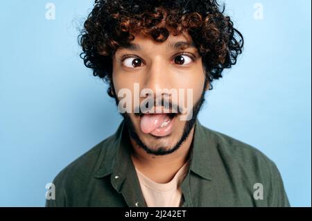 Close up face of indian or arabian curly funny guy, making a funny face, tongue out, squinted his eyes, standing on isolated blue background. Grimaces, fooling around Stock Photo