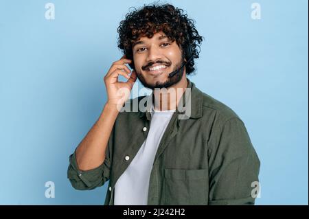 Portrait of friendly positive indian or arabian young adult man, call center, hotline employee with headphones and microphone, stands on isolated blue background, looks at the camera, smiling Stock Photo