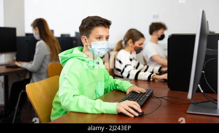 Positive male student wearing face mask working on computer in library. Concept of adult self education during pandemic Stock Photo