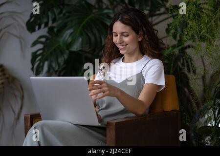 Young happy female gardener sitting on chair with laptop while working remotely at her plant shop Stock Photo