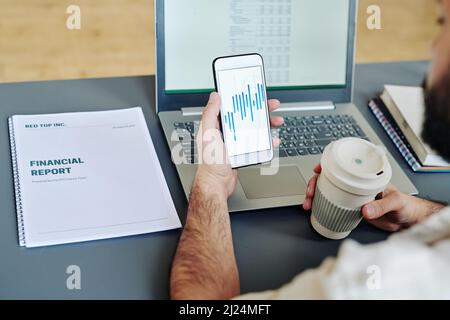 Young male office worker holding glass of coffee and smartphone with financial charts on screen while analyzing data by workplace Stock Photo
