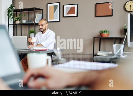 Serious young businessman with mug of tea looking through online data on laptop screen while analyzing it and having drink in office Stock Photo