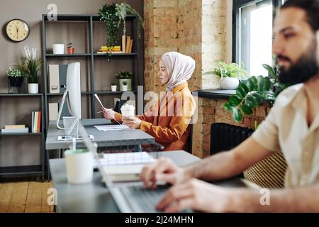 Contemporary Muslim businesswoman analyzing financial data in documents while sitting in front of computer monitor Stock Photo
