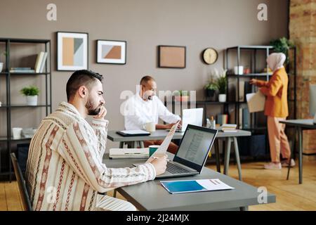Serious young male economist analyzing financial papers in front of laptop by workplace in office against two busy coworkers Stock Photo