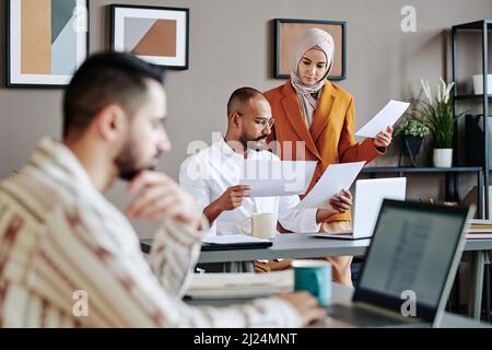 Two young Muslim coworkers looking through financial documents by workplace in front of male broker sitting in front of laptop Stock Photo