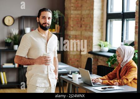 Happy young Muslim man in casual apparel holding cup of coffee in front of himself while standing against female colleague Stock Photo