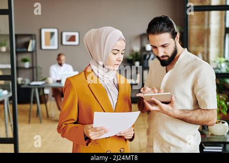 Young businessman scrolling in tablet while standing next to his female colleague with paper during presentation or planning work Stock Photo