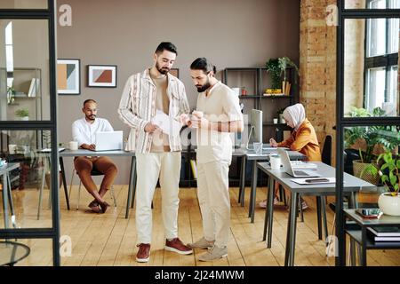 Two young Muslim businessmen in smart casualwear discussing financial documents or contract while standing in the center of office Stock Photo