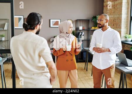 Group of young Muslim coworkers having coffee and discussing plans at break while standing in the center of openspace office Stock Photo