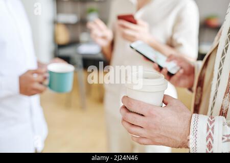 Hand of young businessman holding cup of coffee and looking through new messages in smartphone while standing among colleagues Stock Photo