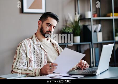 Serious bearded male economist looking through financial document while sitting in front of laptop and analyzing information Stock Photo
