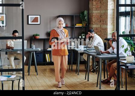 Young serious Muslim businesswoman in hijab reading financial documents while walking along openspace office Stock Photo