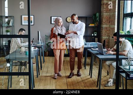 Young confident economist pointing at financial document held by Muslim female colleague while explaining her data Stock Photo