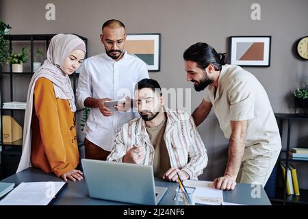 Young confident businessman pointing at data on laptop screen while making presentation for colleagues at working meeting Stock Photo