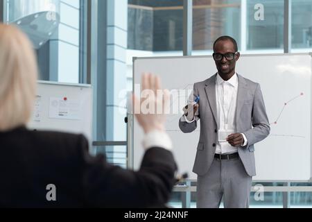 Young smiling African American teacher pointing at female broker raising hand to answer question about presentation at training Stock Photo