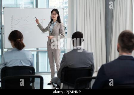 Successful business coach pointing at graphs on whiteboard at seminar while making presentation of fluctuations to colleagues Stock Photo