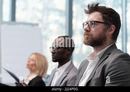 Young serious bearded businessman looking at speaker during presentation against two intercultural colleagues Stock Photo