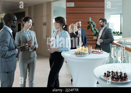Young intercultural entrepreneurs with champagne discussing best reports after conference while standing by served tables Stock Photo