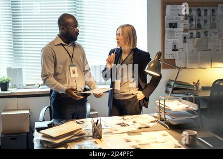 Young serious African American male FBI agent listening to mature female colleague in formalwear with pen by her chin Stock Photo