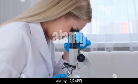 Female laboratory assistant analyzes samples using a microscope while working at a table in a laboratory, doing research Stock Photo
