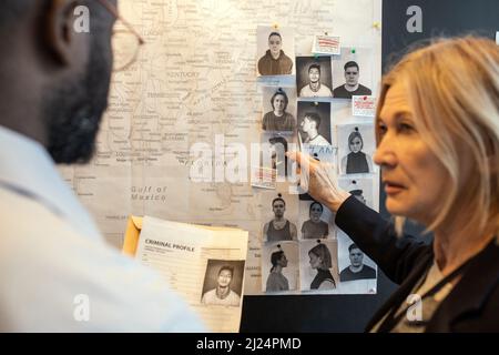 Blond confident female detective pointing at photo of suspect on board over map while matching facts of serial crimes with colleague Stock Photo