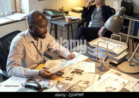 Serious male agent of FBI learning criminal profiles by desk in office while his female colleague speaking to witness on the phone Stock Photo