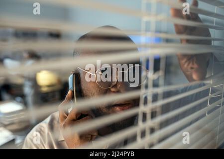 African American male security system department staff talking on the phone while looking through venetian blinds in office Stock Photo