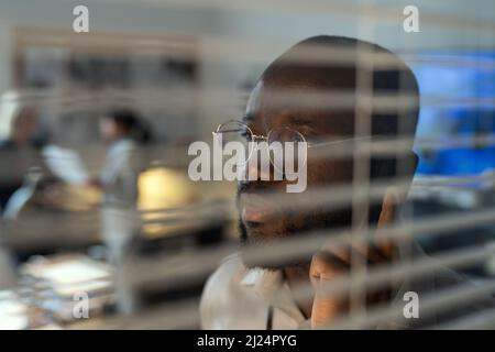 Serious African American FBI agent talking on the phone by venetian blinds while standing in office against two female colleagues Stock Photo