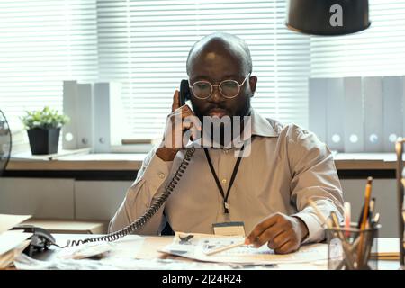 Serious African American FBI agent in formalwear speaking on telephone while sitting by workplace and looking through documents Stock Photo