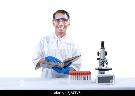 Asian nerd scientist standing and holding a book with a microscope and medical tube rack on the desk isolated over white background Stock Photo