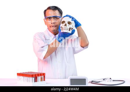 Asian nerd scientist standing and holding a skull head with a stethoscope and medical tube rack on the desk isolated over white background Stock Photo