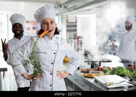 Funny playful head chef having carrot between teeth acting goofy and childish while posing for camera. Amusing gastronomy expert with vegetable in mouth smiling heartily while in restaurant kitchen. Stock Photo