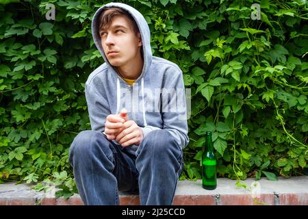 Sad Young Man in a Hoodie with a Beer Bottle on the Sidewalk of the City Street Stock Photo