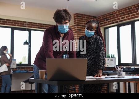 Multi ethnic diverse office workers wearing facemasks against coronavirus analyzing company accounting data and reviewing partner portfolio. Expectant mother finance advisor looking over project ideas Stock Photo