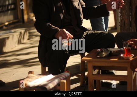 Elderly men drinking tea holding prayer beads in traditional village coffee sitting on wooden stool on sidewalk. Stock Photo