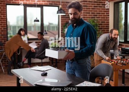 Sad dismissed man holding box with belongings after being fired while his collegues looking at hom working in startup office. Businesspeople brainstorming ideas developing marketing project Stock Photo
