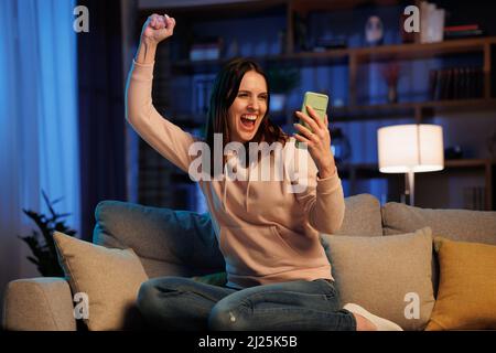 Happy young woman surprised by the good shocking and unbelievable news, making yes gesture while looking at smartphone. Enjoying success, celebrating Stock Photo
