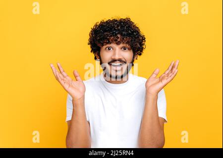 Surprised confused indian or arabian guy with curly hair, wearing white t-shirt, looking at camera in disbelief, spreading his arms to the sides, shrugging, standing over isolated orange background Stock Photo